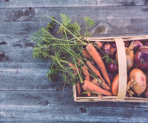 carrots and onions in a wood basket ontop of a wooden table.